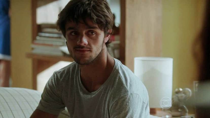 Jonatas está sentado em um sofá em cena de Totalmente Demais com camisa cinza e entre luz e sombra iluminando seu rosto e corpo Jonatas está sentado em um sofá em cena de Totalmente Demais com camisa cinza e entre luz e sombra iluminando seu rosto e corpo