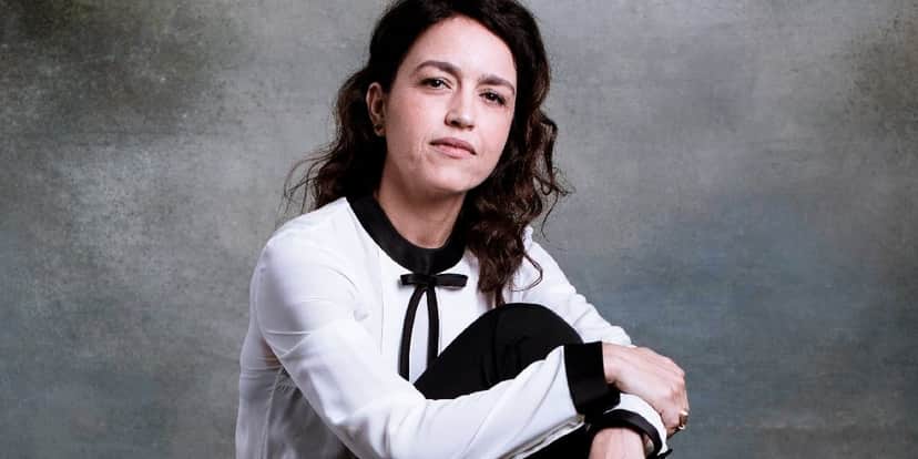 Manuela Dias sob um fundo cinza, é uma mulher branca, de cabelos cacheados castanhos que está sentada segurando sua perna com as mãos, usa roupas em tons brancos e pretos Manuela Dias sob um fundo cinza, é uma mulher branca, de cabelos cacheados castanhos que está sentada segurando sua perna com as mãos, usa roupas em tons brancos e pretos