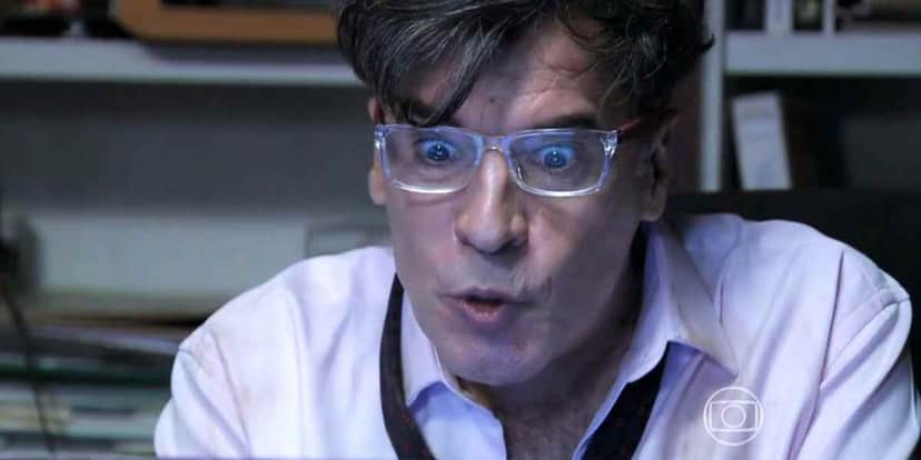 Teo está em cena de Império de frente a uma tela, usa óculos transparentes, camisa branca e gravata preta Teo está em cena de Império de frente a uma tela, usa óculos transparentes, camisa branca e gravata preta