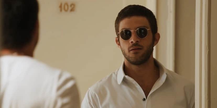 Ari usando camisa branca e óculos escuro parado em frente a porta aberta na novela Travessia Ari usando camisa branca e óculos escuro parado em frente a porta aberta na novela Travessia