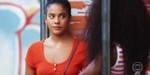 Jeniffer da novela Vai na Fé é uma mulher jovem, negra que está usando uma camiseta laranja e o cabelo está preso Jeniffer da novela Vai na Fé é uma mulher jovem, negra que está usando uma camiseta laranja e o cabelo está preso