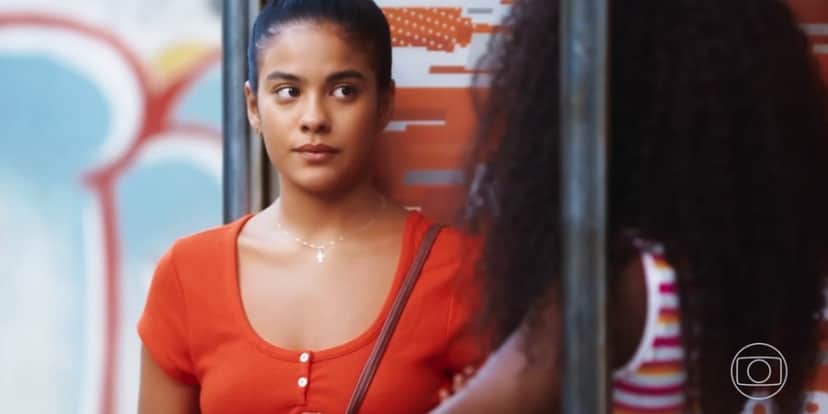 Jeniffer da novela Vai na Fé é uma mulher jovem, negra que está usando uma camiseta laranja e o cabelo está preso Jeniffer da novela Vai na Fé é uma mulher jovem, negra que está usando uma camiseta laranja e o cabelo está preso