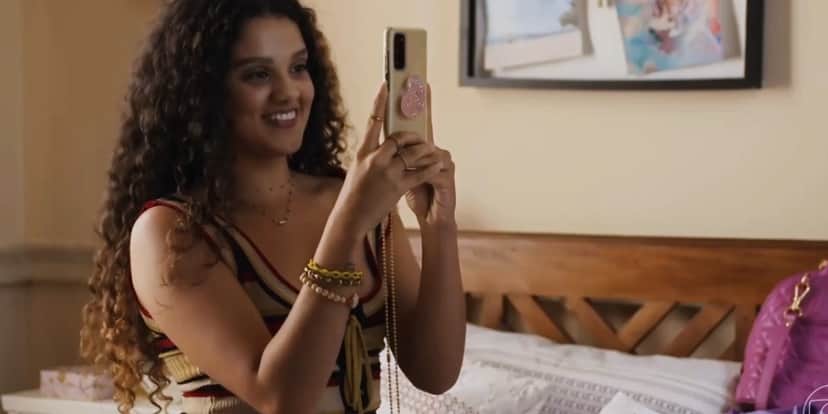 Na trama de Travessia Karina está usando uma regata listrada, cabelo longo cacheado e segura um celular na mão Na trama de Travessia Karina está usando uma regata listrada, cabelo longo cacheado e segura um celular na mão