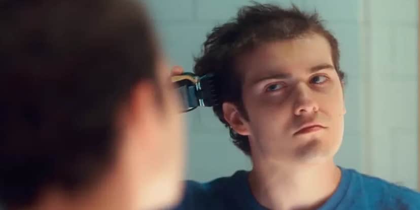 Rafa personagem de Vai na Fé usa camiseta azul e se olha no espelho para raspar o cabelo