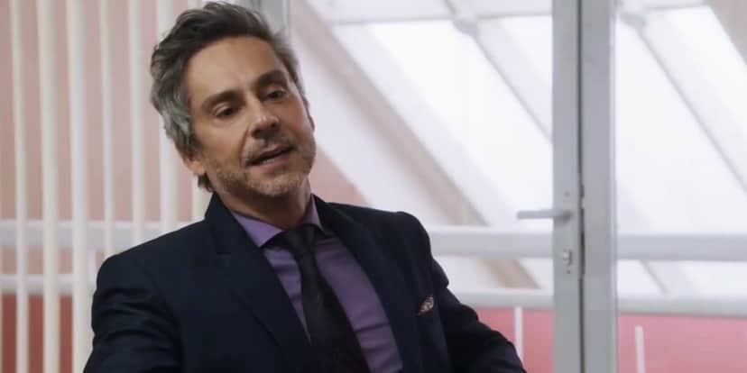 Stenio sentado em cena da novela Travessia, usando terno azul escuro, gravata vinho e camisa social roxa
