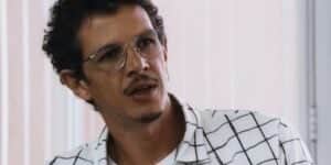 Gil em cena da novela Travessia usando camisa xadrez branca e preta, óculos de grau prata e cabelo curto com cavanhaque preto Gil em cena da novela Travessia usando camisa xadrez branca e preta, óculos de grau prata e cabelo curto com cavanhaque preto
