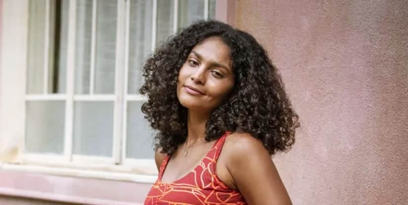 Bárbara Reis vestindo um vestido vermelho com listras brancas, com cabelo curto cacheado castanho escuro em bastidor de Terra e Paixão Bárbara Reis vestindo um vestido vermelho com listras brancas, com cabelo curto cacheado castanho escuro em bastidor de Terra e Paixão