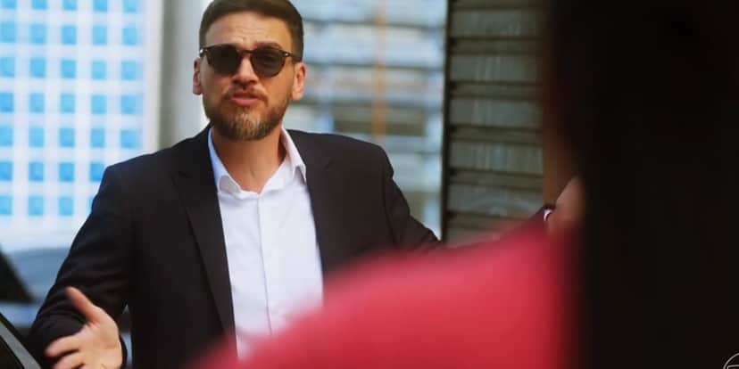 Theo na rua usando terno azul escuro aberto, camisa branca e óculos escuros em cena da novela Vai na Fé Theo na rua usando terno azul escuro aberto, camisa branca e óculos escuros em cena da novela Vai na Fé