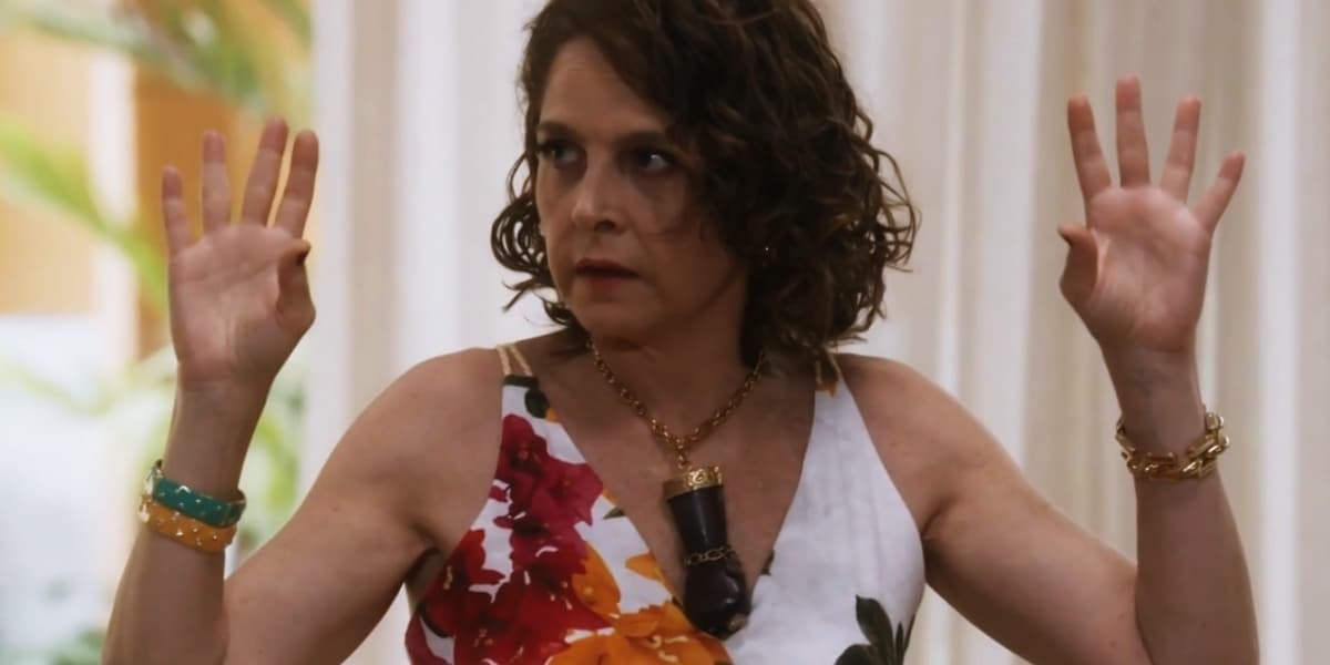 Núbia em cena da novela Travessia usando vestido florido e colar com grande mão preta com punho fechado Núbia em cena da novela Travessia usando vestido florido e colar com grande mão preta com punho fechado