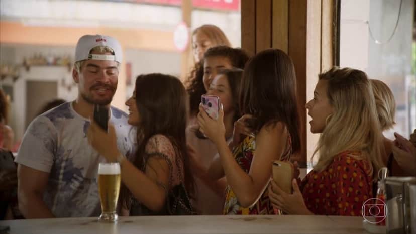 Em cena da novela Amor de Mãe, o ator Thiago Martins como o personagem Ryan está em um bar sendo abordado por algumas garotas com celular na mão Em cena da novela Amor de Mãe, o ator Thiago Martins como o personagem Ryan está em um bar sendo abordado por algumas garotas com celular na mão