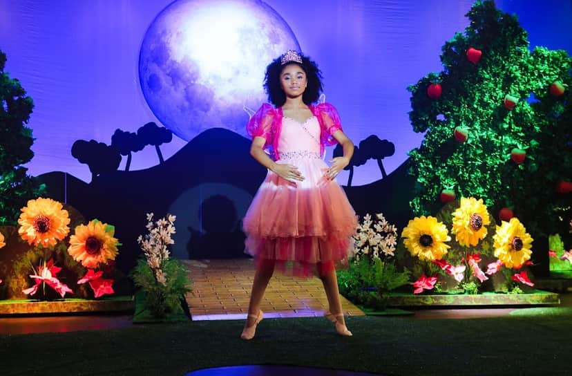 Cenário colorido de uma peça de teatro onde Kessya de As Aventuras de Poliana está no centro em pose de bailarina e vestido rosa Cenário colorido de uma peça de teatro onde Kessya de As Aventuras de Poliana está no centro em pose de bailarina e vestido rosa