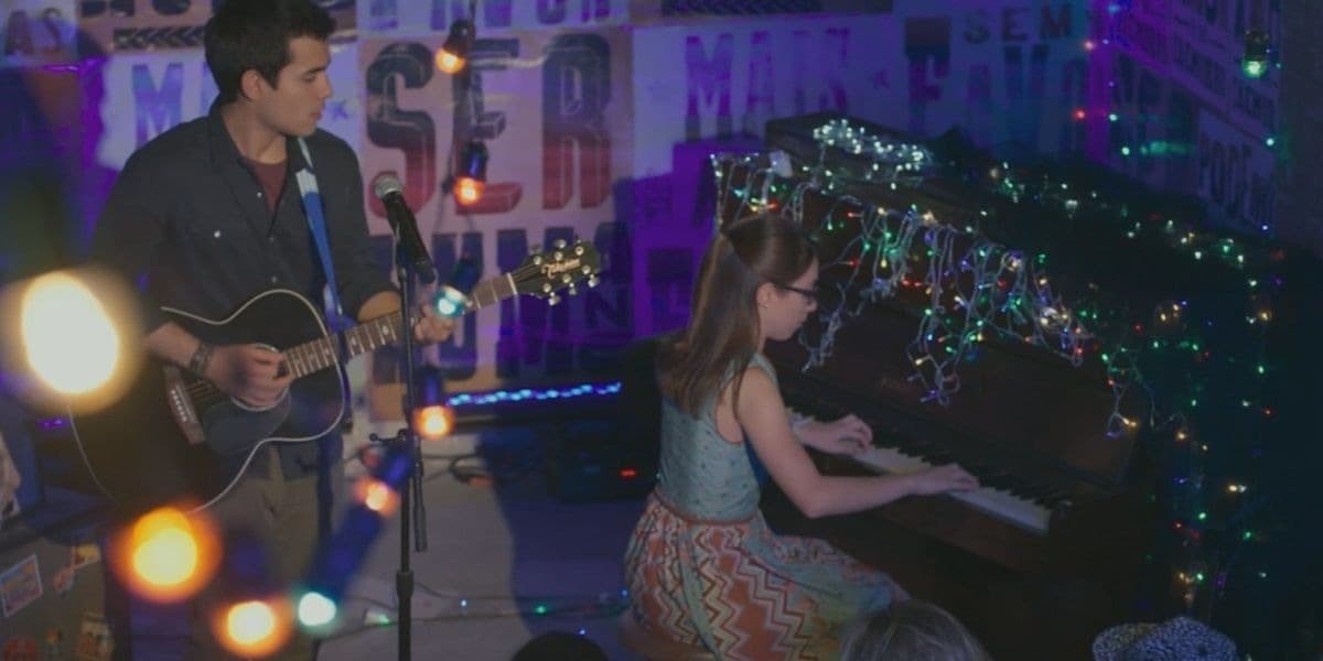 Na foto da cena da novela Malhação - Viva a Diferença em ambiente escuro iluminado por diversas luzes de natal, Benê esta no piano e Guto em pé cantando e tocando em frente a plateia Na foto da cena da novela Malhação - Viva a Diferença em ambiente escuro iluminado por diversas luzes de natal, Benê esta no piano e Guto em pé cantando e tocando em frente a plateia