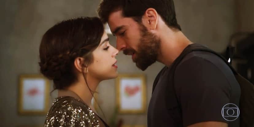 Em cena da novela Salve-se Quem Puder Micaela com casaco com brilhoso e bruno com camisa azul escura estão frente a frente prestes a se beijarem Em cena da novela Salve-se Quem Puder Micaela com casaco com brilhoso e bruno com camisa azul escura estão frente a frente prestes a se beijarem
