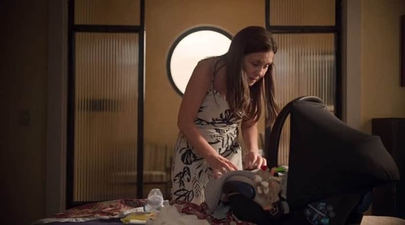 Thelma em cena de Amor de Mãe, mulher branca, com cabelos longos lisos e vestido florido está abaixada de frente ao bebê conforto em um quarto Thelma em cena de Amor de Mãe, mulher branca, com cabelos longos lisos e vestido florido está abaixada de frente ao bebê conforto em um quarto