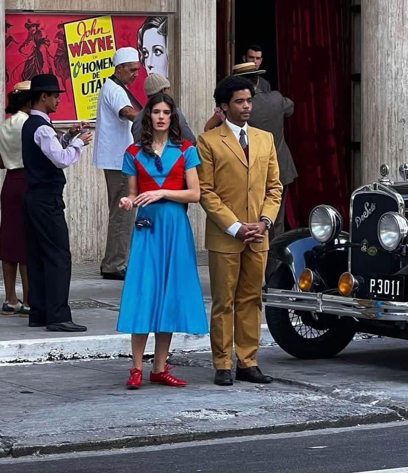 Atores Camila Queiroz e Diogo Almeida estão no rua perto de um carro preto antigo ela usa vestido longo azul com vermelho e ele está vestido com terno e calça amarelo escuro Atores Camila Queiroz e Diogo Almeida estão no rua perto de um carro preto antigo ela usa vestido longo azul com vermelho e ele está vestido com terno e calça amarelo escuro