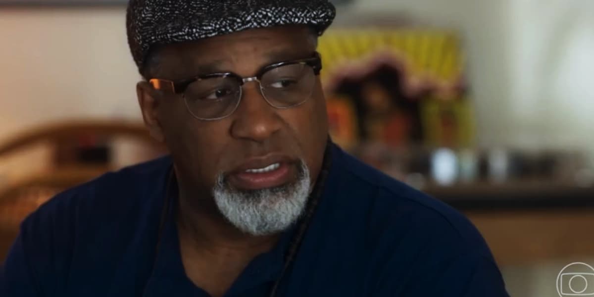Monteiro usando camisa azul, boina xadrez e colar de bolinhas pretas em cena da novela Travessia Monteiro usando camisa azul, boina xadrez e colar de bolinhas pretas em cena da novela Travessia