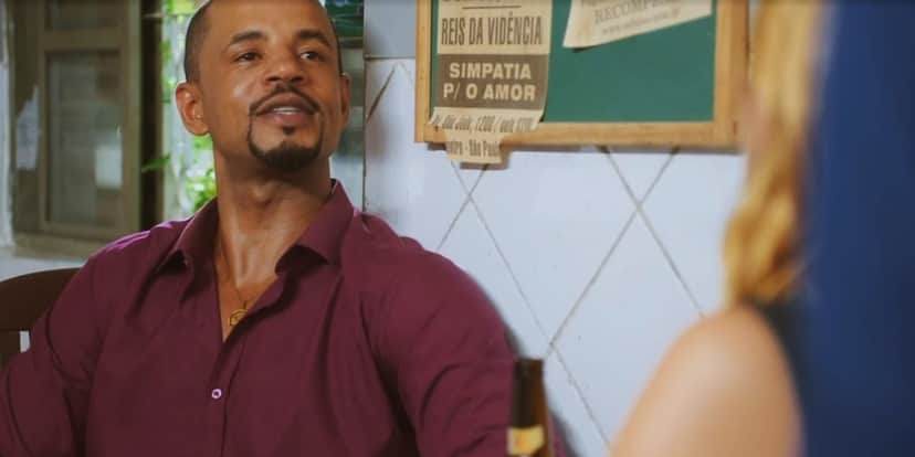 Orfeu está sentado em mesa de bar usando camisa vinho em Vai na Fé Orfeu está sentado em mesa de bar usando camisa vinho em Vai na Fé